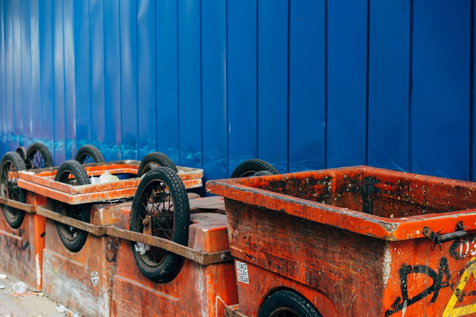 Image showing a row of orange and black waste containers positioned against a tall blue metal fence. The containers are rectangular with open tops and exhibit signs of wear, dirt, and some graffiti pa