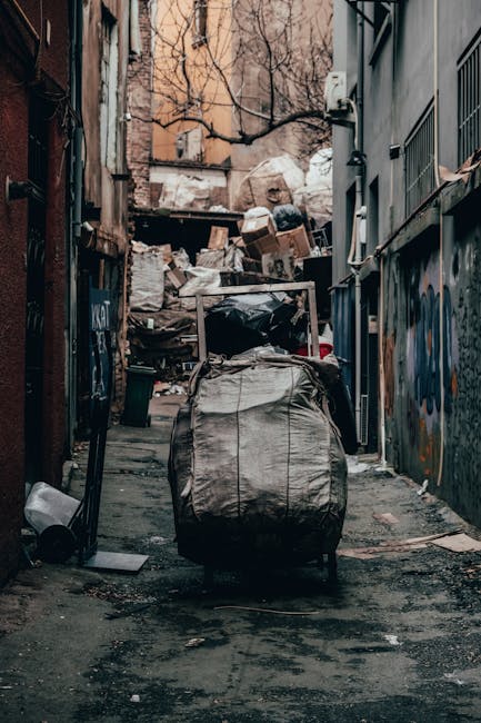 A person wearing a yellow and gray plaid shirt, gray trousers, and bright green gloves is holding open a large, black trash bag made of glossy plastic material, preparing for rubbish disposal. The ind