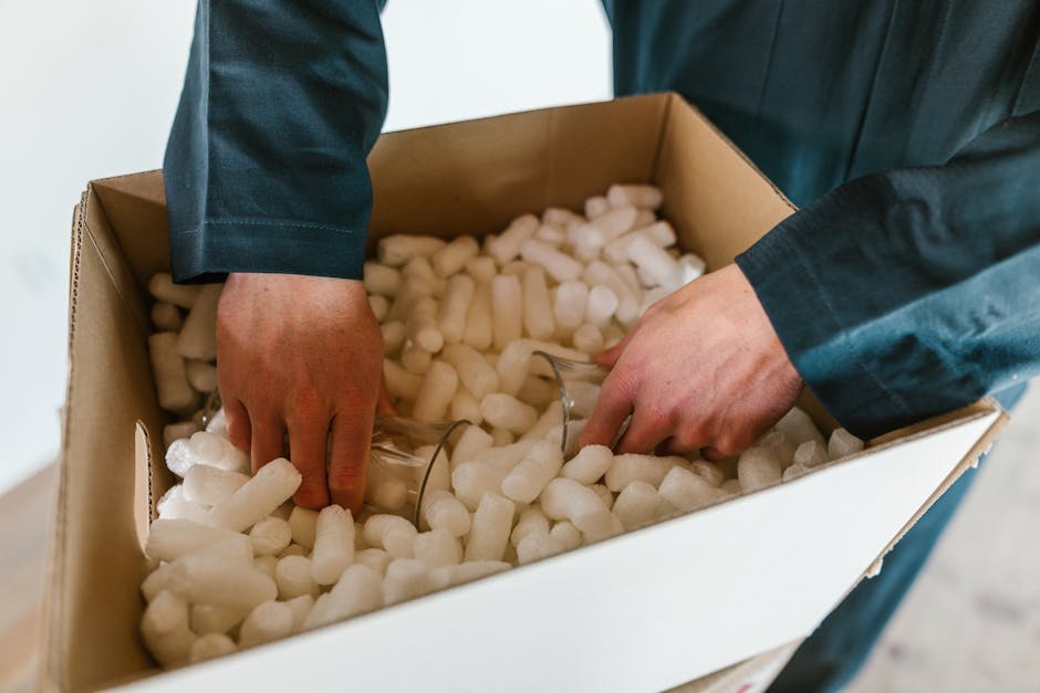 A person is carefully wrapping a fragile, green ceramic or glass object in layers of brown kraft paper to protect it during handling, with their hands gently securing the packing material around the i