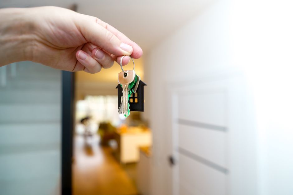 A close-up of a person's hand holding a keyring with multiple keys, including a brass key, a green plastic key tag, and a miniature house-shaped key fob, positioned in the foreground. The background s