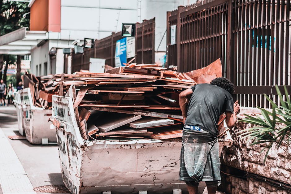 A narrow urban alleyway cluttered with overflowing waste and discarded items, including large black and white garbage bags, cardboard boxes, and miscellaneous debris piled against a back wall. The gro