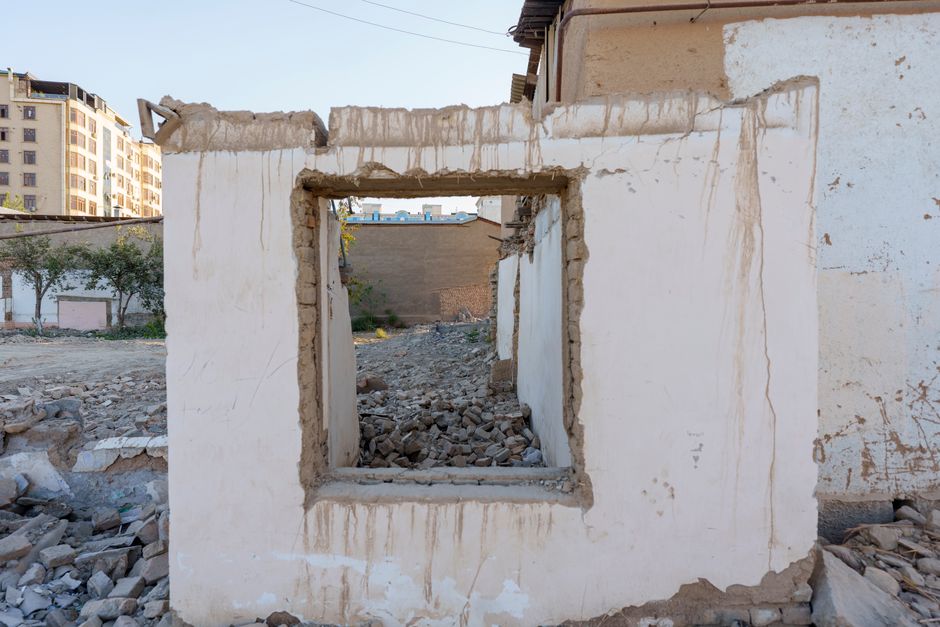 A partially demolished section of a white plastered wall with visible brick and concrete edges, featuring a rectangular window opening with rough, uneven borders. Debris, including small stones, concr