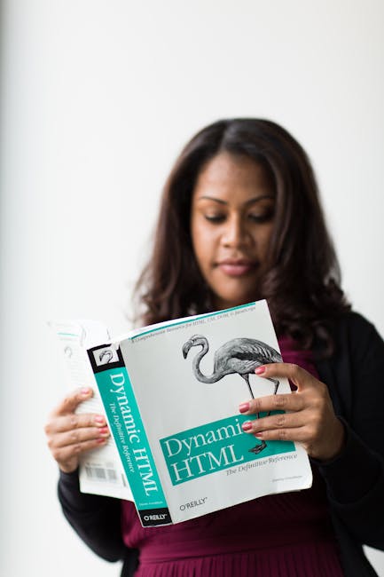 A woman with medium brown skin and long, dark brown hair is holding and looking at a book titled 'Dynamic HTML' with a flamingo illustration on the cover, in an indoor setting with plain white walls.